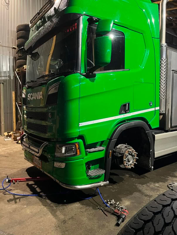 A green truck in a garage with its front wheel removed, tools and a spare tire are visible on the ground nearby.
