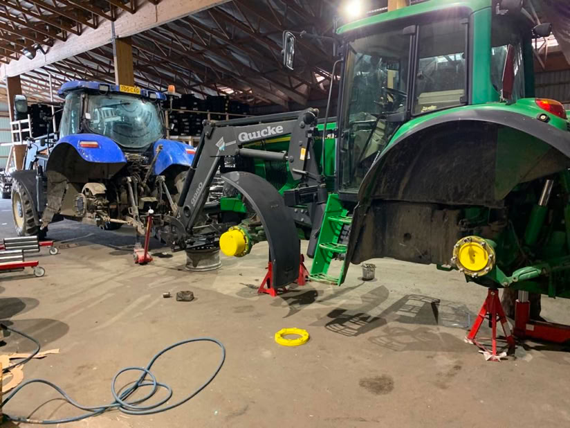 Two tractors, one blue and one green, are parked inside a garage. Both tractors are elevated on red jack stands with their front wheels removed. Various tools and equipment are scattered on the floor.