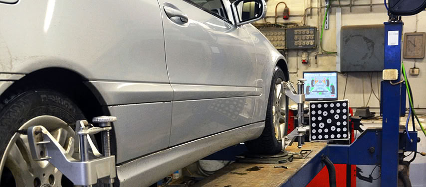 A silver car undergoing wheel alignment in a garage, with equipment attached to the wheels and a computer screen displaying data.