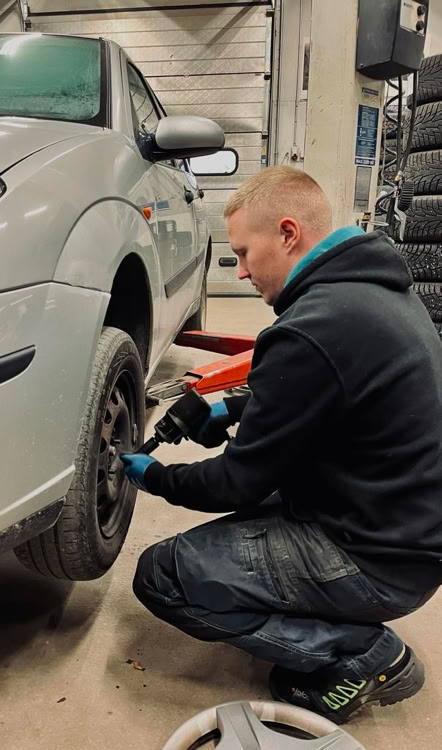 A person kneeling and working on the front wheel of a silver car in a garage setting.