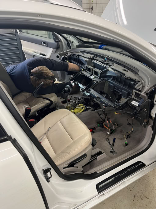 A person working inside a car with the dashboard removed, exposing wires and components.